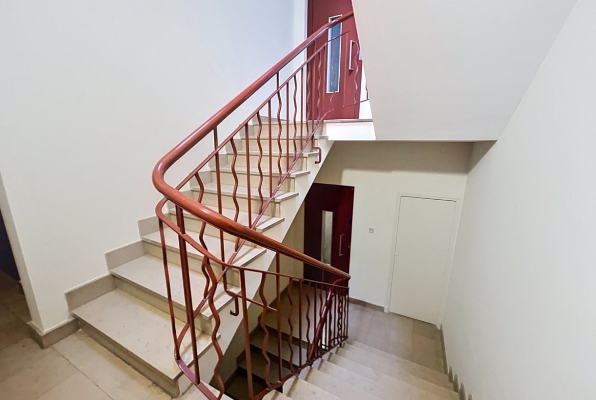Escalier en pierre beige avec rampe métallique rouge à motifs ondulés et main courante en bois. Murs blancs, portes d'appartement rouge bordeaux aux étages, sol en carrelage clair dans le hall.