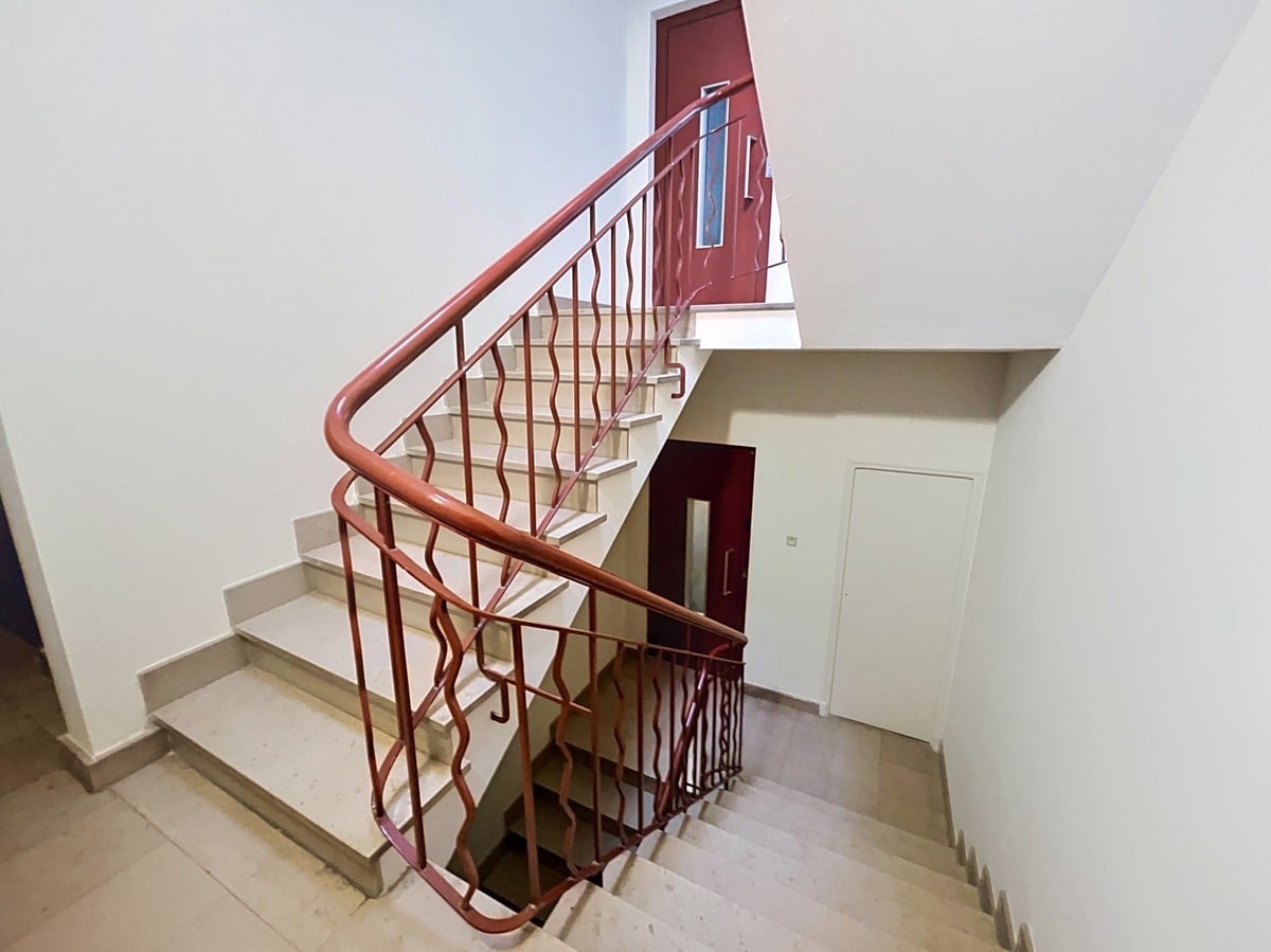 Escalier en pierre beige avec rampe métallique rouge à motifs ondulés et main courante en bois. Murs blancs, portes d'appartement rouge bordeaux aux étages, sol en carrelage clair dans le hall.