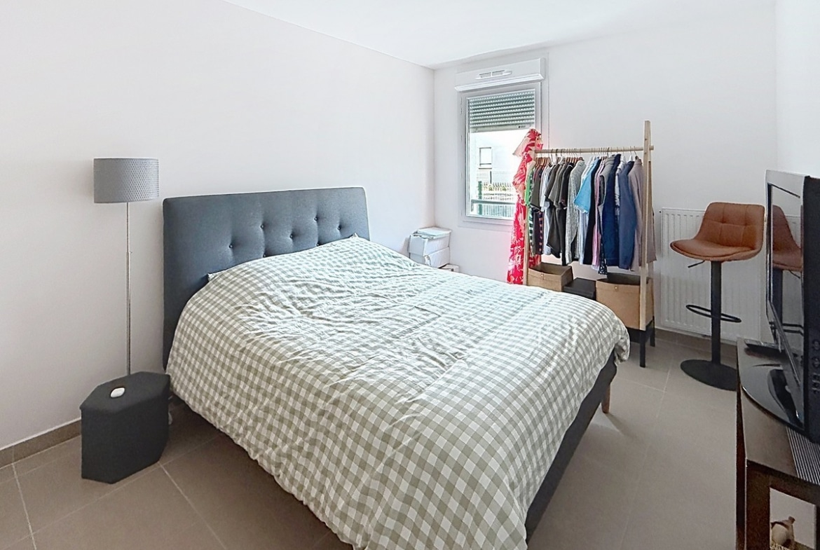 Chambre avec lit double doté d'une tête de lit grise capitonnée et couette à carreaux gris et blanc. Un portant en bois avec vêtements colorés est installé près de la fenêtre, accompagné d'un lampadaire gris et d'une chaise haute en cuir marron.