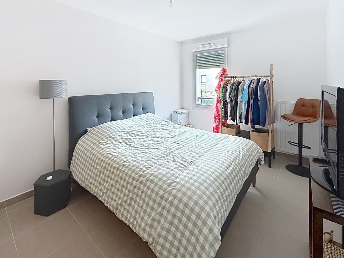 Chambre avec lit double doté d'une tête de lit grise capitonnée et couette à carreaux gris et blanc. Un portant en bois avec vêtements colorés est installé près de la fenêtre, accompagné d'un lampadaire gris et d'une chaise haute en cuir marron.