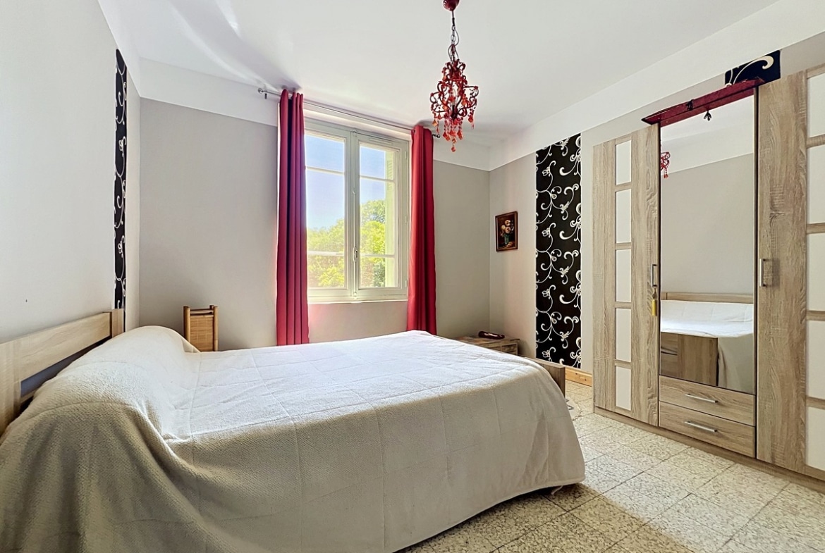 Chambre aux murs gris clair et blanc avec lit double recouvert d'un couvre-lit beige. Armoire en bois clair avec porte miroir, rideaux roses encadrant une fenêtre et lustre rouge suspendu au plafond blanc.