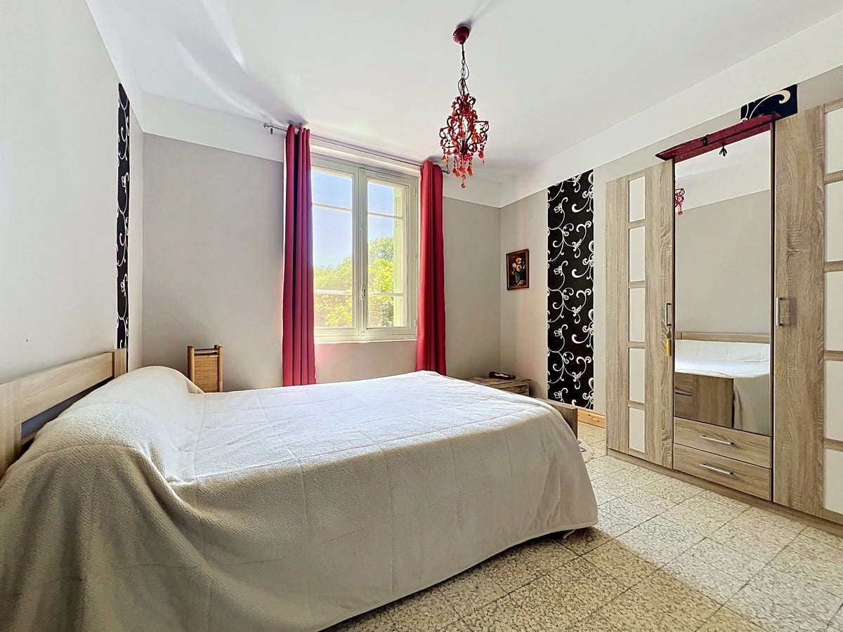 Chambre aux murs gris clair et blanc avec lit double recouvert d'un couvre-lit beige. Armoire en bois clair avec porte miroir, rideaux roses encadrant une fenêtre et lustre rouge suspendu au plafond blanc.