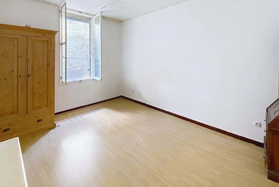 Chambre aux murs blancs avec parquet stratifié clair et plinthes en carrelage bordeaux. Une armoire en pin massif est installée près de la fenêtre à battants qui donne sur un mur en pierre, et un ancien poêle à bois en fonte est visible dans l'angle.