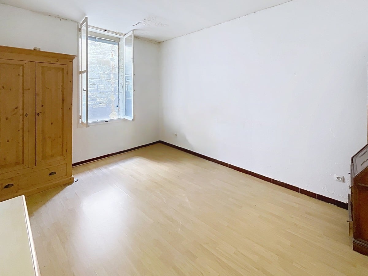 Chambre aux murs blancs avec parquet stratifié clair et plinthes en carrelage bordeaux. Une armoire en pin massif est installée près de la fenêtre à battants qui donne sur un mur en pierre, et un ancien poêle à bois en fonte est visible dans l'angle.
