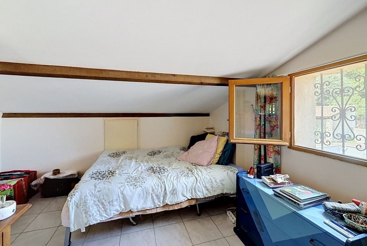 Chambre mansardée avec poutre apparente en bois et plafond en pente blanc. Lit double avec couette blanche, fenêtre en bois avec grille en fer forgé et commode bleue.