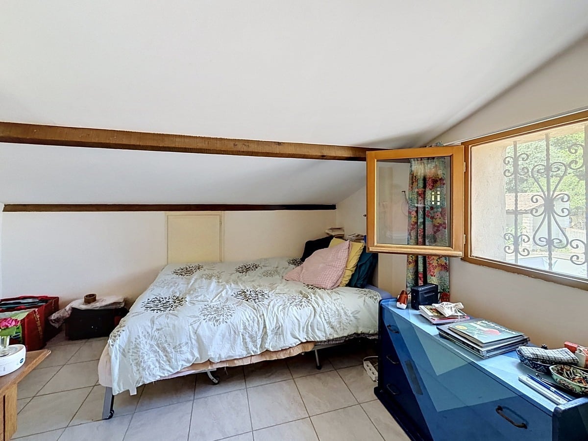 Chambre mansardée avec poutre apparente en bois et plafond en pente blanc. Lit double avec couette blanche, fenêtre en bois avec grille en fer forgé et commode bleue.