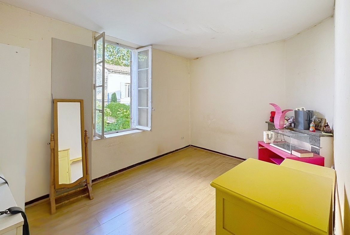Chambre avec parquet stratifié clair, fenêtre à deux battants ouverte sur la verdure et une commode jaune au premier plan. Un miroir sur pied en bois est posé contre le mur gauche et une cheminée décorative avec tablette en marbre gris occupe l'angle droit.