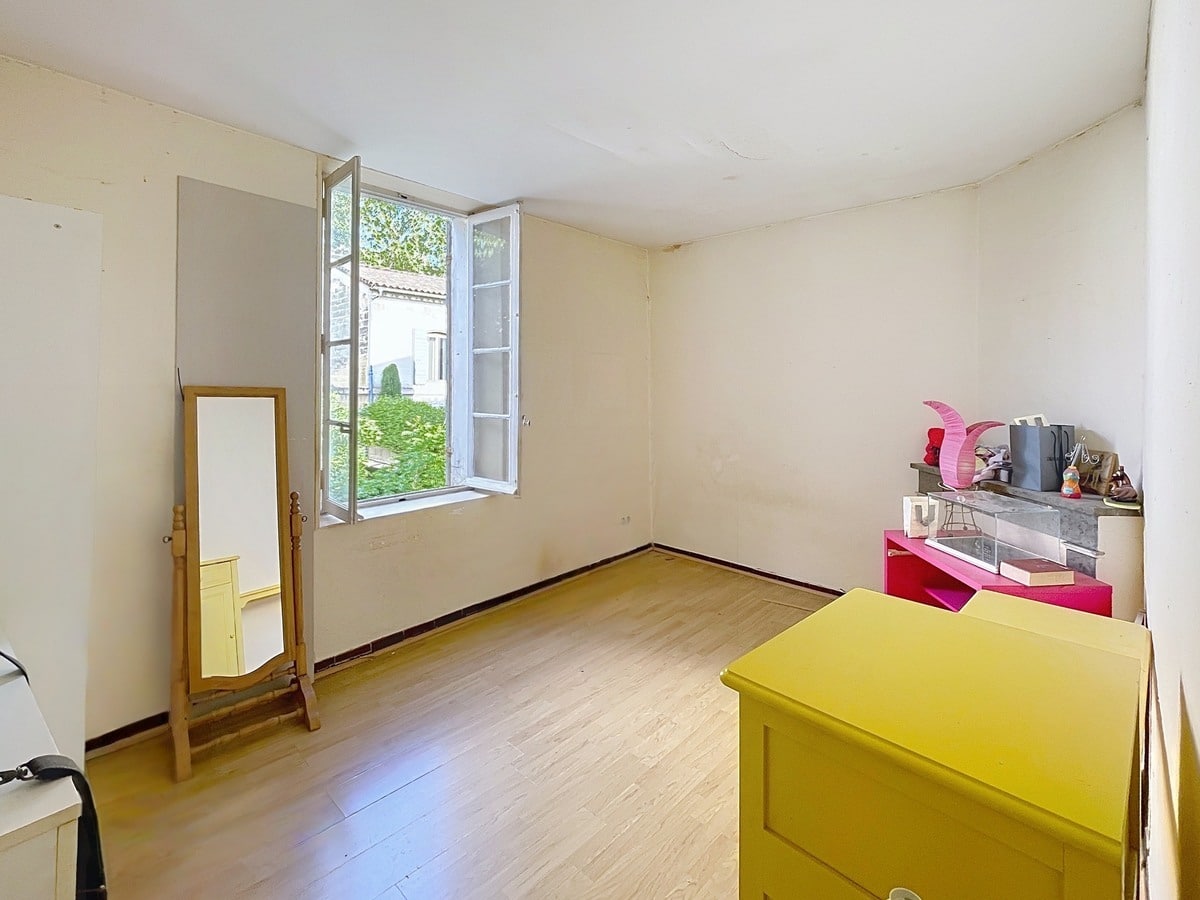 Chambre avec parquet stratifié clair, fenêtre à deux battants ouverte sur la verdure et une commode jaune au premier plan. Un miroir sur pied en bois est posé contre le mur gauche et une cheminée décorative avec tablette en marbre gris occupe l'angle droit.