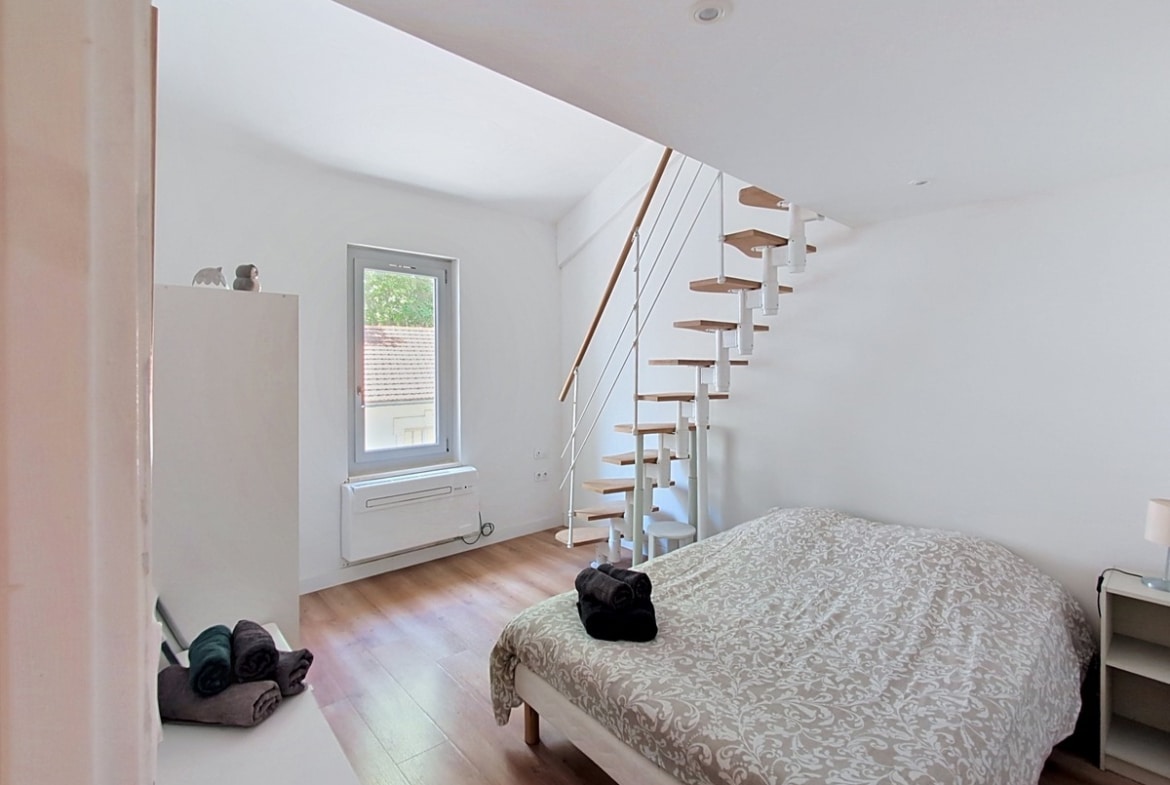 Chambre aux murs blancs avec parquet stratifié clair et lit double recouvert d'une couette à motifs beiges. Un escalier à pas japonais en bois et métal blanc mène à une mezzanine, avec une fenêtre à cadre gris donnant sur des toits en tuiles.