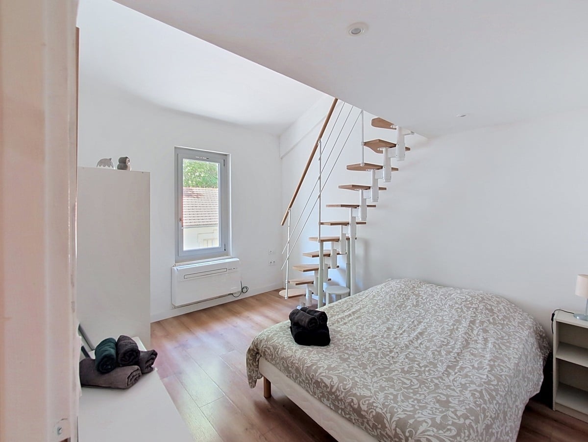 Chambre aux murs blancs avec parquet stratifié clair et lit double recouvert d'une couette à motifs beiges. Un escalier à pas japonais en bois et métal blanc mène à une mezzanine, avec une fenêtre à cadre gris donnant sur des toits en tuiles.