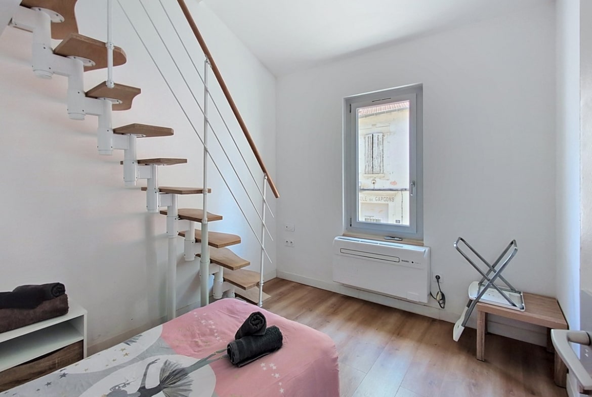 Chambre blanche avec lit simple recouvert d'une housse rose à motif étoilé. Un escalier à pas japonais en bois et métal blanc longe le mur. La fenêtre PVC grise révèle une ancienne inscription "École de Garçons" sur le bâtiment voisin.