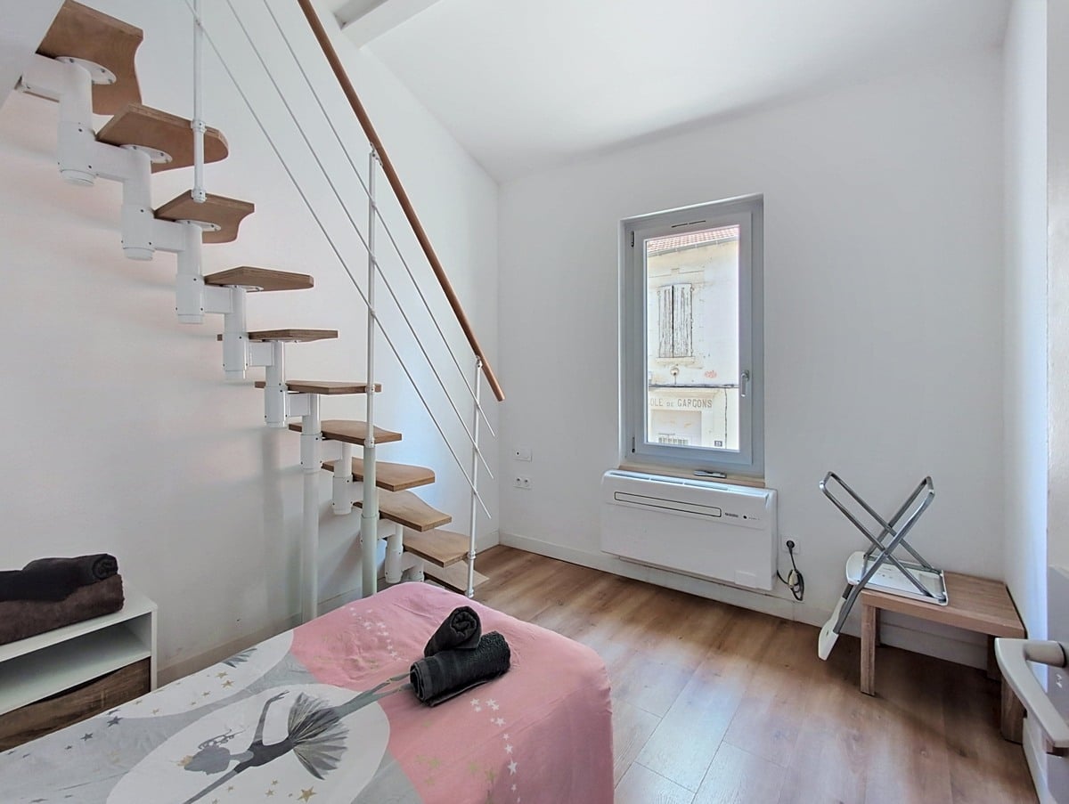 Chambre blanche avec lit simple recouvert d'une housse rose à motif étoilé. Un escalier à pas japonais en bois et métal blanc longe le mur. La fenêtre PVC grise révèle une ancienne inscription "École de Garçons" sur le bâtiment voisin.
