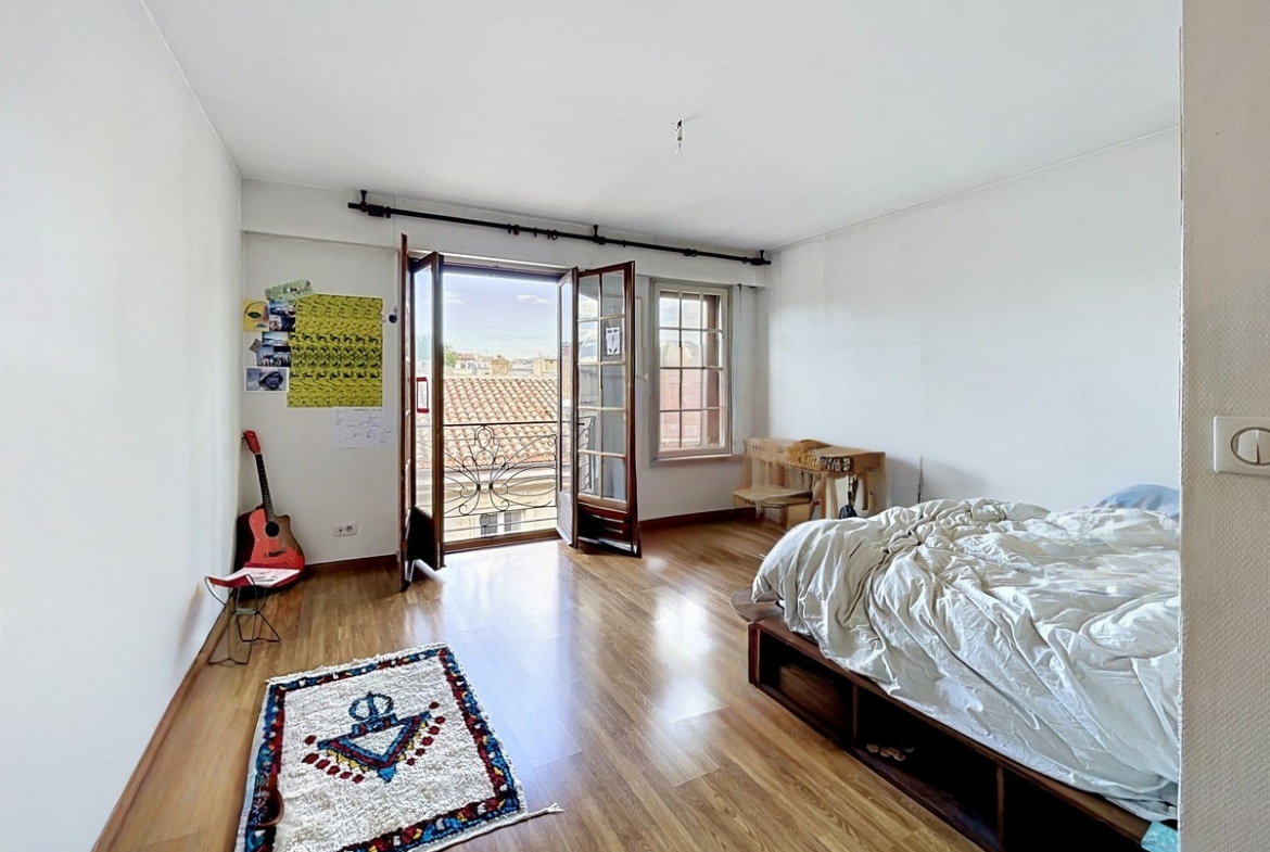 Chambre avec lit sur cadre en bois à rangements intégrés, parquet clair au sol et tapis berbère à motif ancre. Portes-fenêtres en bois ouvrant sur un balcon avec garde-corps en fer forgé, vue sur toits en tuiles, guitare rouge posée contre le mur blanc.