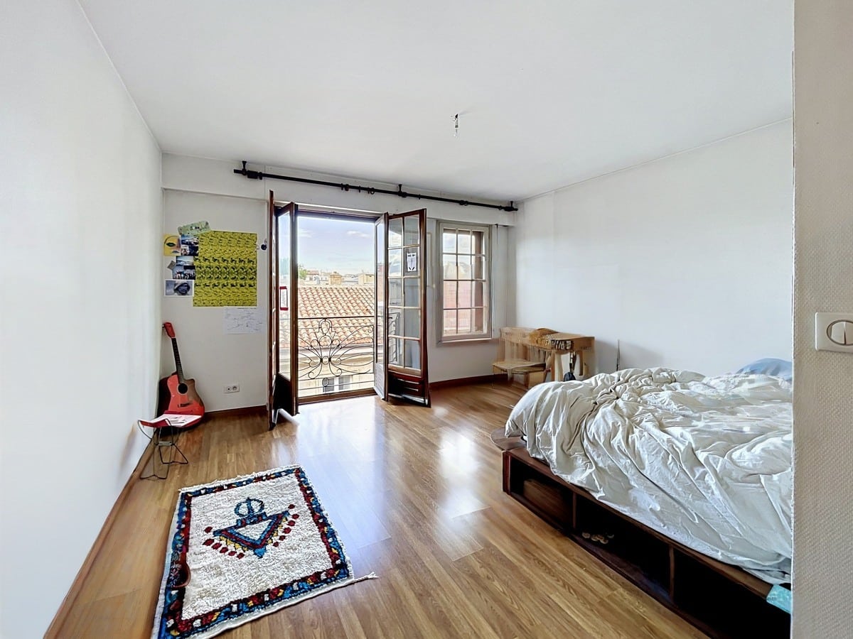 Chambre avec lit sur cadre en bois à rangements intégrés, parquet clair au sol et tapis berbère à motif ancre. Portes-fenêtres en bois ouvrant sur un balcon avec garde-corps en fer forgé, vue sur toits en tuiles, guitare rouge posée contre le mur blanc.