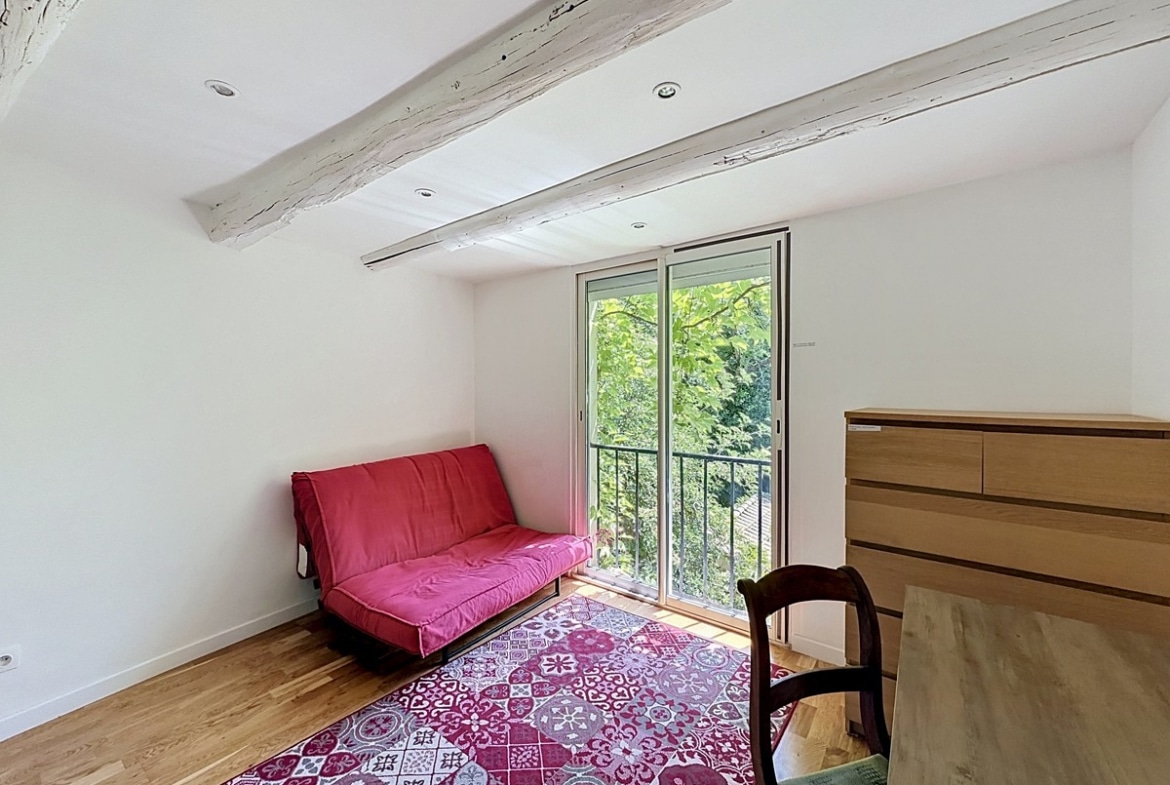 Chambre sous combles avec deux poutres en bois peintes en blanc, parquet clair et porte-fenêtre donnant sur verdure. Canapé-lit rouge, tapis à motifs carreaux de ciment bordeaux, commode en bois clair et chaise ancienne.