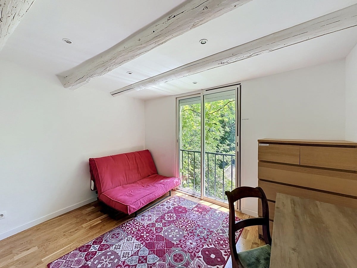 Chambre sous combles avec deux poutres en bois peintes en blanc, parquet clair et porte-fenêtre donnant sur verdure. Canapé-lit rouge, tapis à motifs carreaux de ciment bordeaux, commode en bois clair et chaise ancienne.