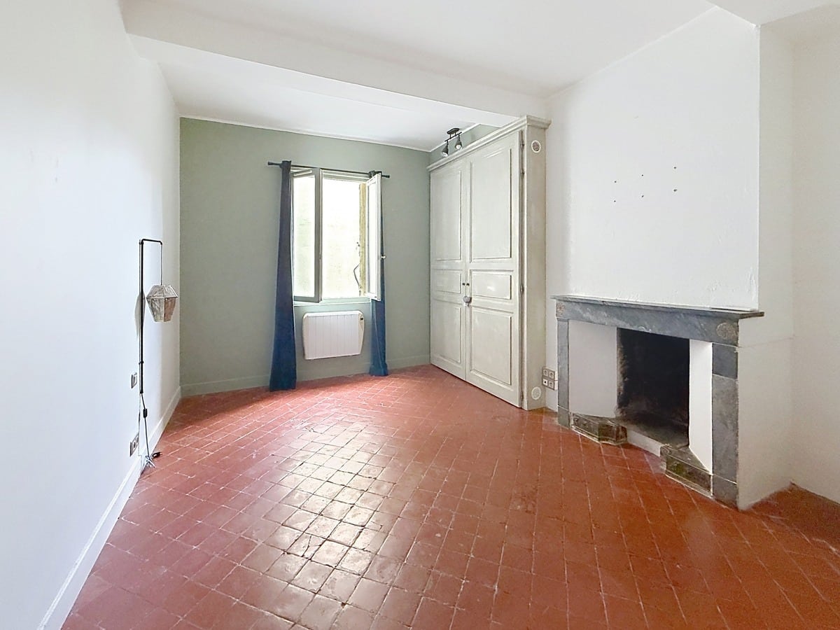 Pièce avec sol en tomettes rouges traditionnelles et cheminée ancienne en marbre gris. Un placard intégré blanc est encastré près de la fenêtre avec rideaux bleu marine, mur d'accent vert sauge.