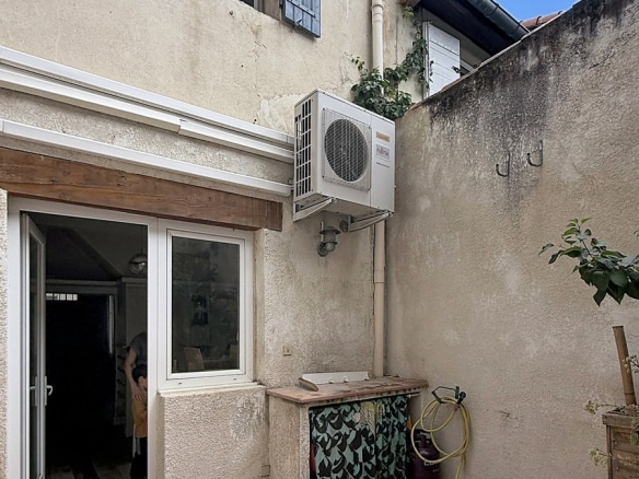 Cour intérieure avec façade ancienne crépi beige et volets bleus à l'étage. Une unité de climatisation est fixée au mur au-dessus d'une porte-fenêtre coulissante blanche donnant sur l'intérieur.