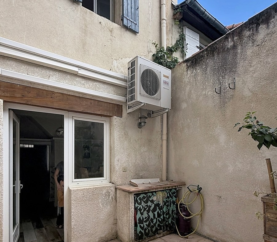 Cour intérieure avec façade ancienne crépi beige et volets bleus à l'étage. Une unité de climatisation est fixée au mur au-dessus d'une porte-fenêtre coulissante blanche donnant sur l'intérieur.
