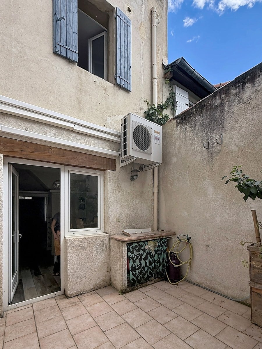 Cour intérieure avec façade ancienne crépi beige et volets bleus à l'étage. Une unité de climatisation est fixée au mur au-dessus d'une porte-fenêtre coulissante blanche donnant sur l'intérieur.