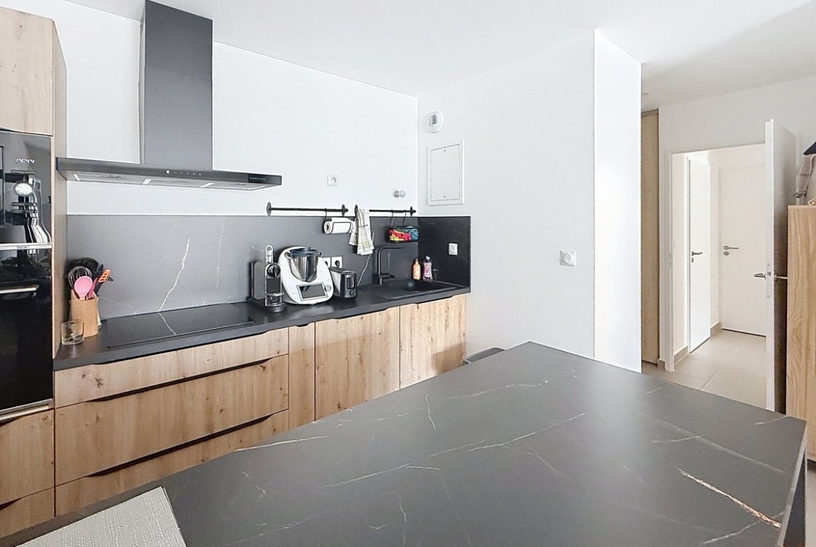 Cuisine avec meubles bas en bois clair et plan de travail noir effet marbre. Îlot central assorti, hotte aspirante noire, robot Thermomix et machine à café visibles sur le plan de travail.