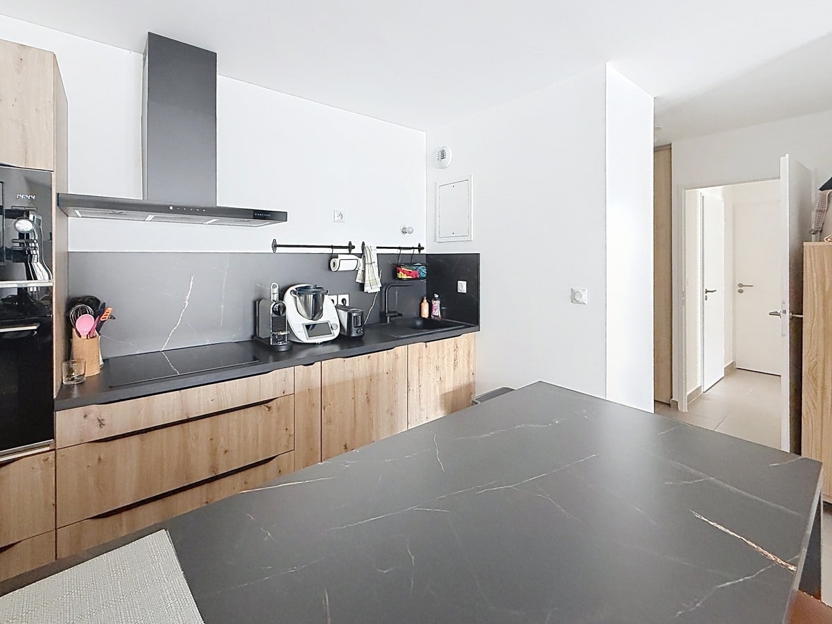 Cuisine avec meubles bas en bois clair et plan de travail noir effet marbre. Îlot central assorti, hotte aspirante noire, robot Thermomix et machine à café visibles sur le plan de travail.