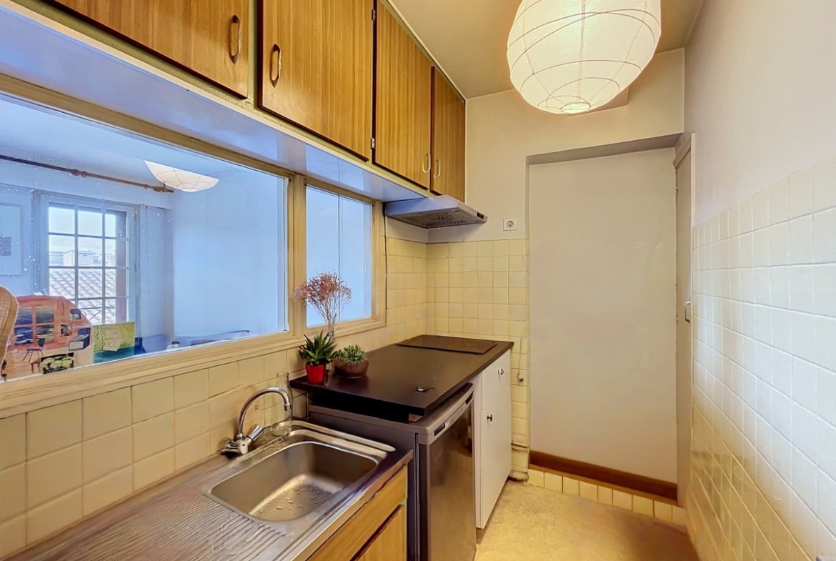 Cuisine étroite avec meubles hauts et bas en bois clair, évier inox et plaque de cuisson noire. Carrelage mural beige, suspension en papier blanc, fenêtre passe-plat donnant sur le séjour, plantes vertes sur le plan de travail.