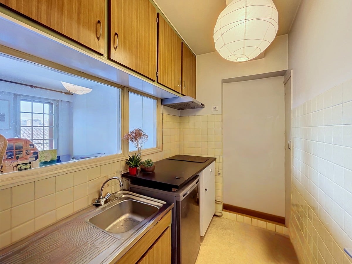 Cuisine étroite avec meubles hauts et bas en bois clair, évier inox et plaque de cuisson noire. Carrelage mural beige, suspension en papier blanc, fenêtre passe-plat donnant sur le séjour, plantes vertes sur le plan de travail.