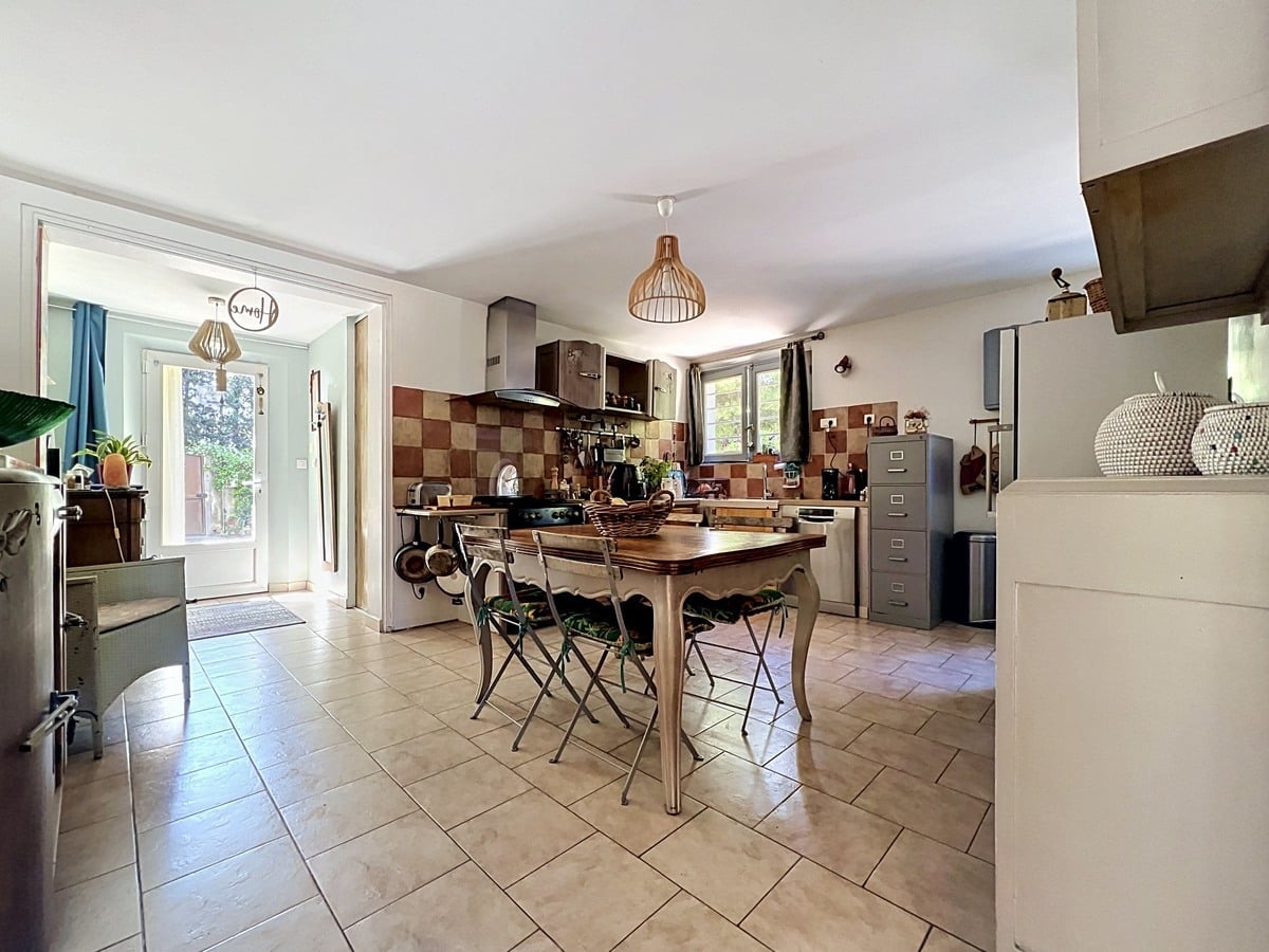 Cuisine avec meubles en bois foncé, crédence en carrelage damier beige et terre cuite, et table ancienne en bois avec chaises pliantes. Un réfrigérateur gris, une hotte aspirante et un classeur métallique sont visibles contre les murs.