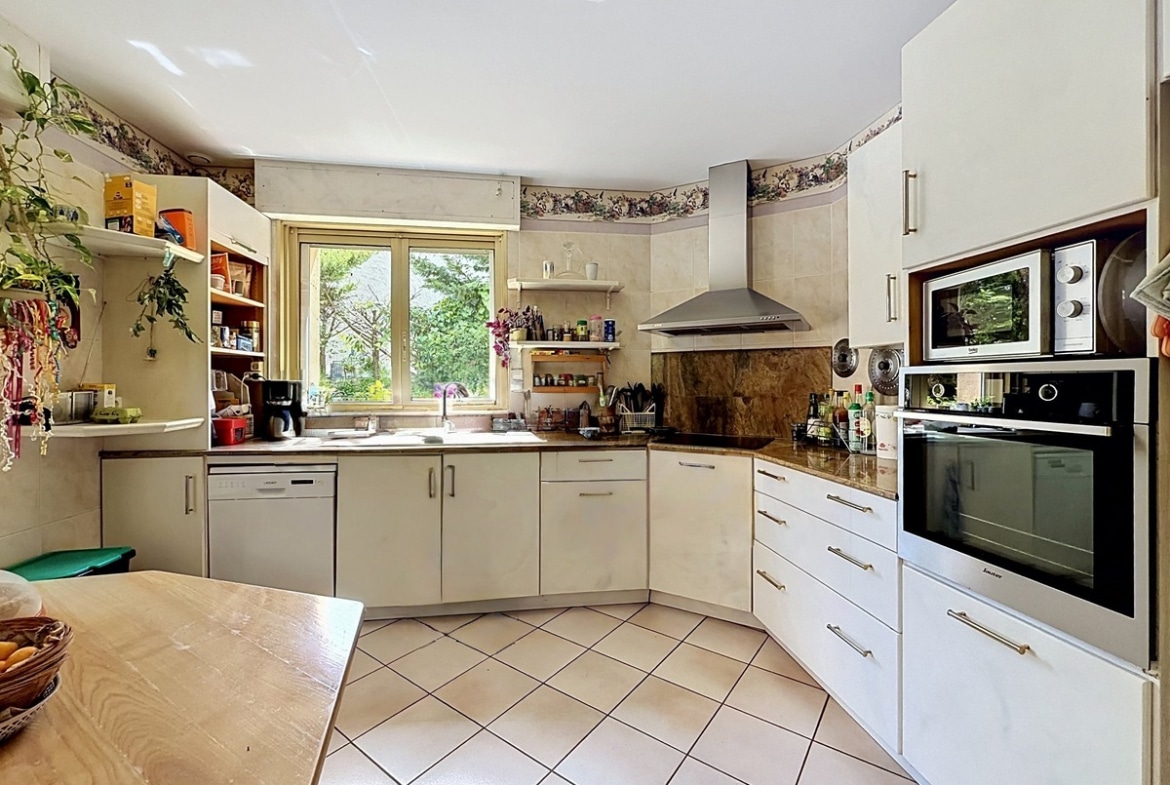 Cuisine en L avec meubles blancs, plan de travail en pierre marbrée et électroménager encastré. Fenêtre donnant sur le jardin, frise décorative florale et îlot central en bois clair.