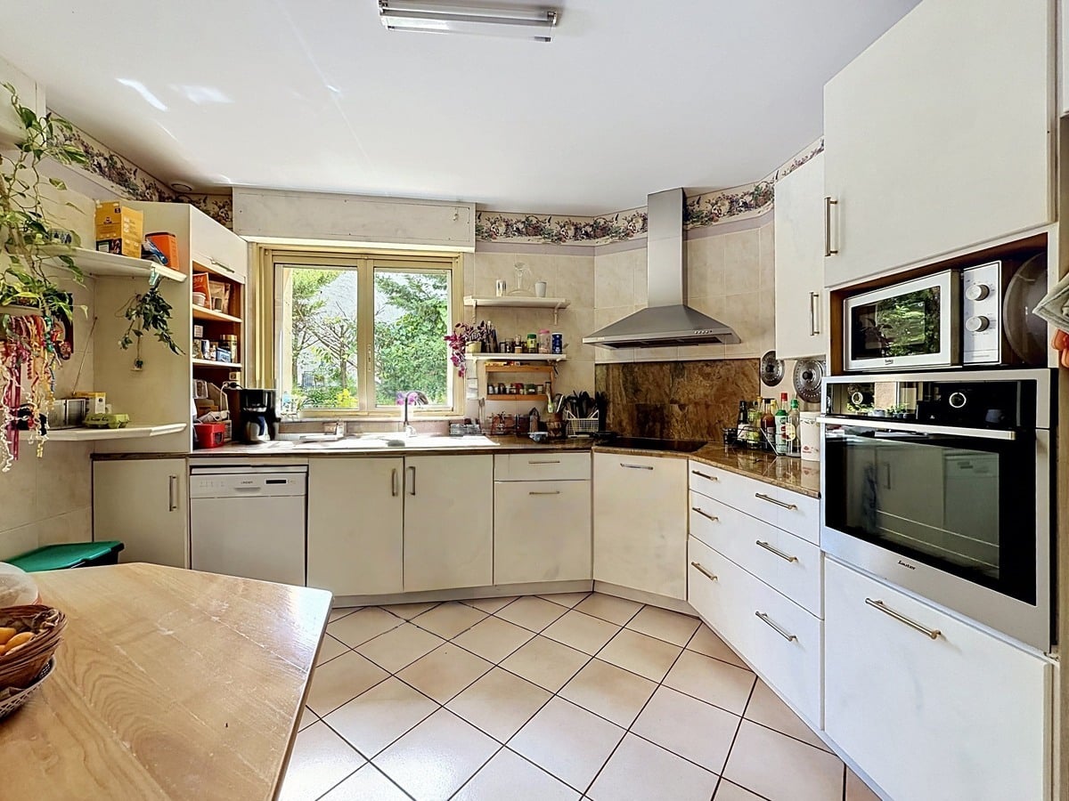Cuisine en L avec meubles blancs, plan de travail en pierre marbrée et électroménager encastré. Fenêtre donnant sur le jardin, frise décorative florale et îlot central en bois clair.