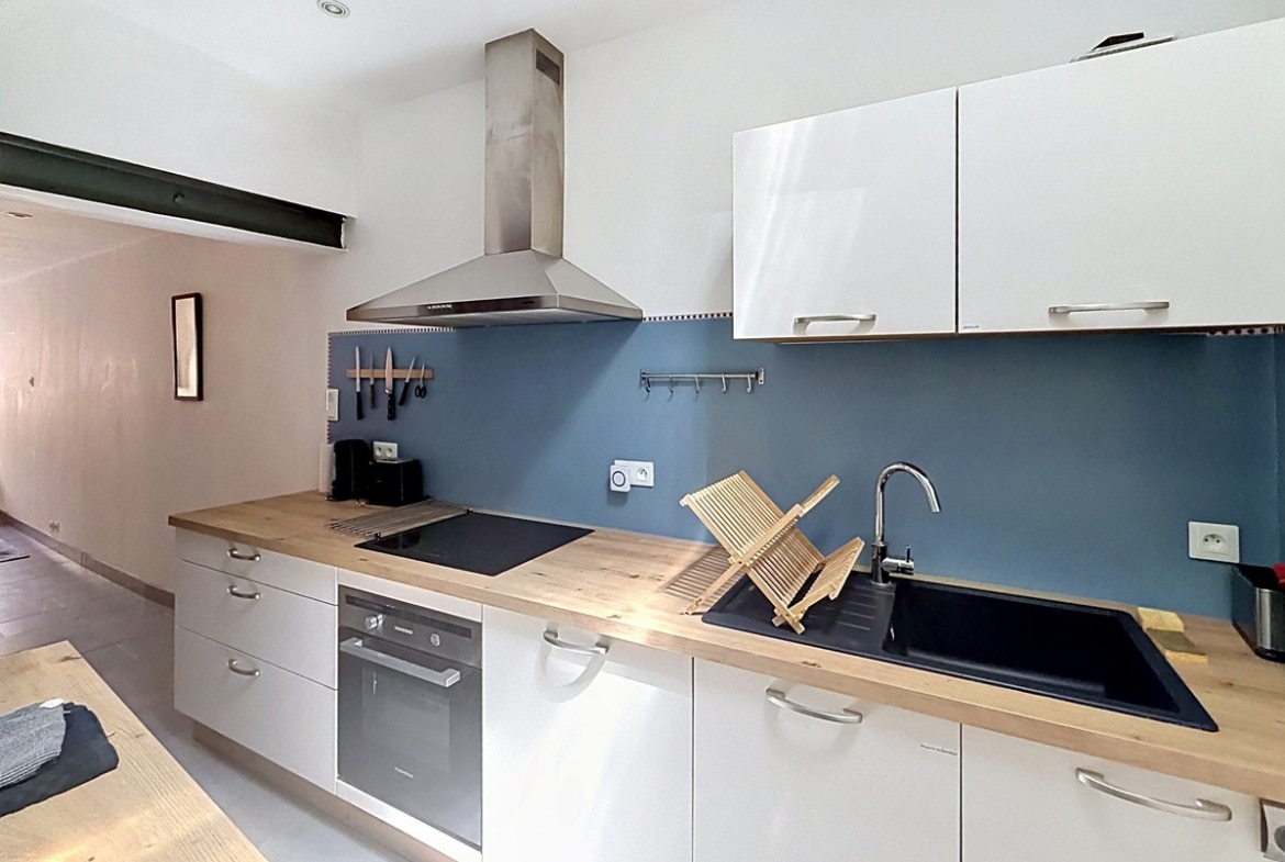 Cuisine linéaire avec meubles blancs et plan de travail en bois massif, crédence bleu-gris. Équipements visibles : plaque à induction, four encastré, hotte en inox, évier noir et égouttoir en bois.