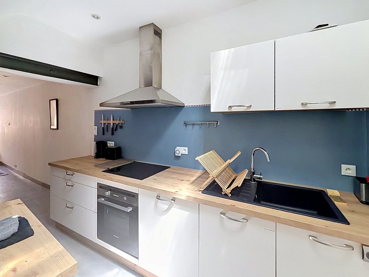 Cuisine linéaire avec meubles blancs et plan de travail en bois massif, crédence bleu-gris. Équipements visibles : plaque à induction, four encastré, hotte en inox, évier noir et égouttoir en bois.