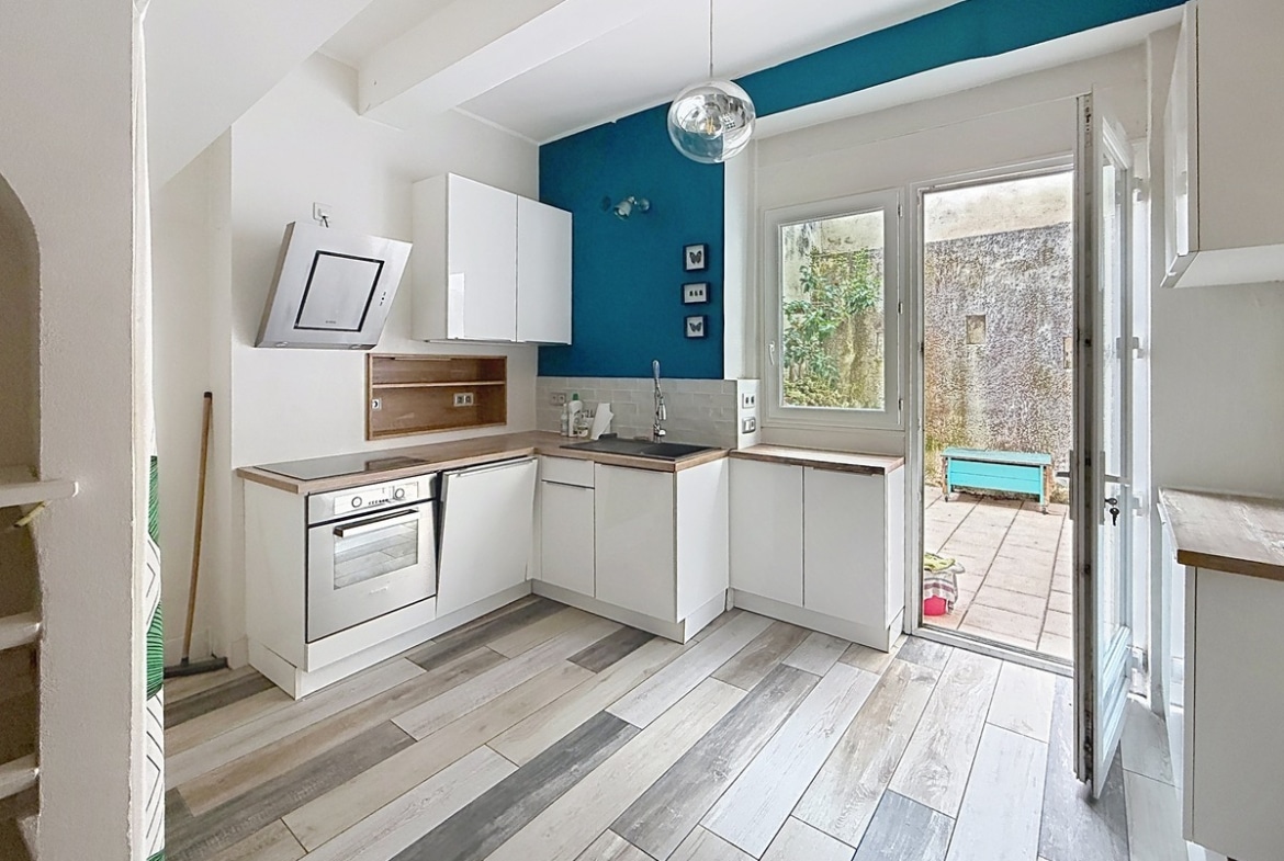 Cuisine ouverte avec meubles blancs et plan de travail en bois, équipée d'un four encastré et d'une hotte inclinée. Le mur d'accent bleu canard contraste avec le sol stratifié gris, une porte-fenêtre ouvre sur la terrasse.