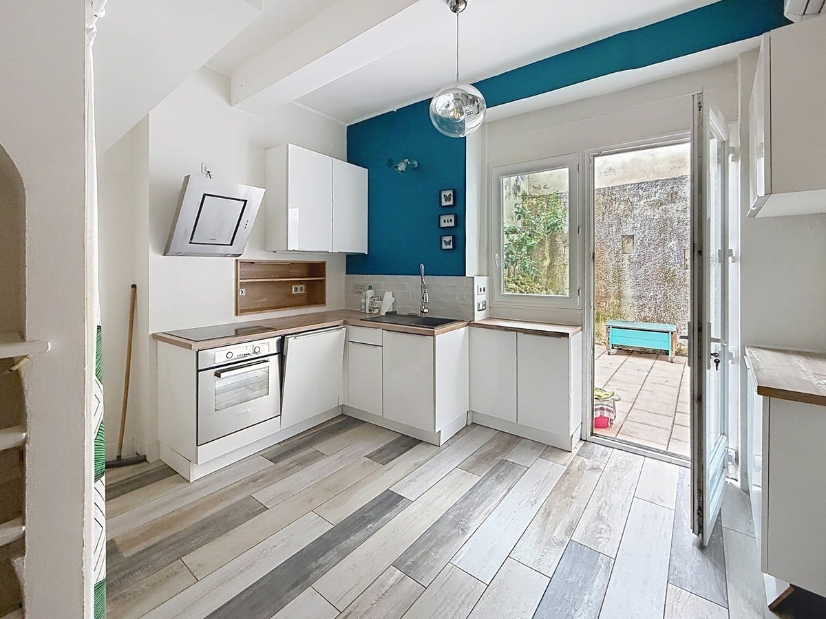 Cuisine ouverte avec meubles blancs et plan de travail en bois, équipée d'un four encastré et d'une hotte inclinée. Le mur d'accent bleu canard contraste avec le sol stratifié gris, une porte-fenêtre ouvre sur la terrasse.