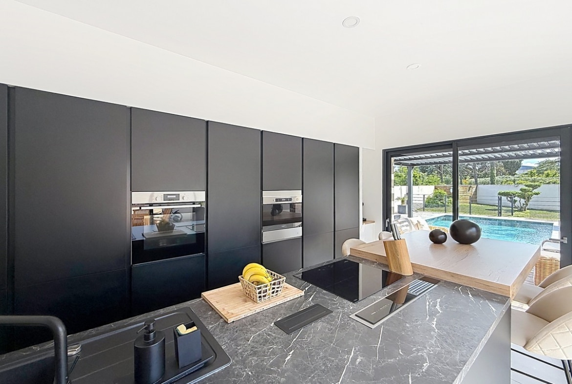 Cuisine contemporaine avec façades noir mat et plan de travail effet marbre gris veiné. Colonne d'électroménagers encastrés et vue directe sur la piscine par baie vitrée coulissante.
