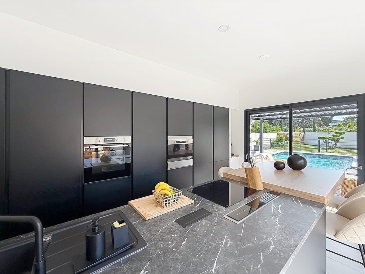Cuisine contemporaine avec façades noir mat et plan de travail effet marbre gris veiné. Colonne d'électroménagers encastrés et vue directe sur la piscine par baie vitrée coulissante.