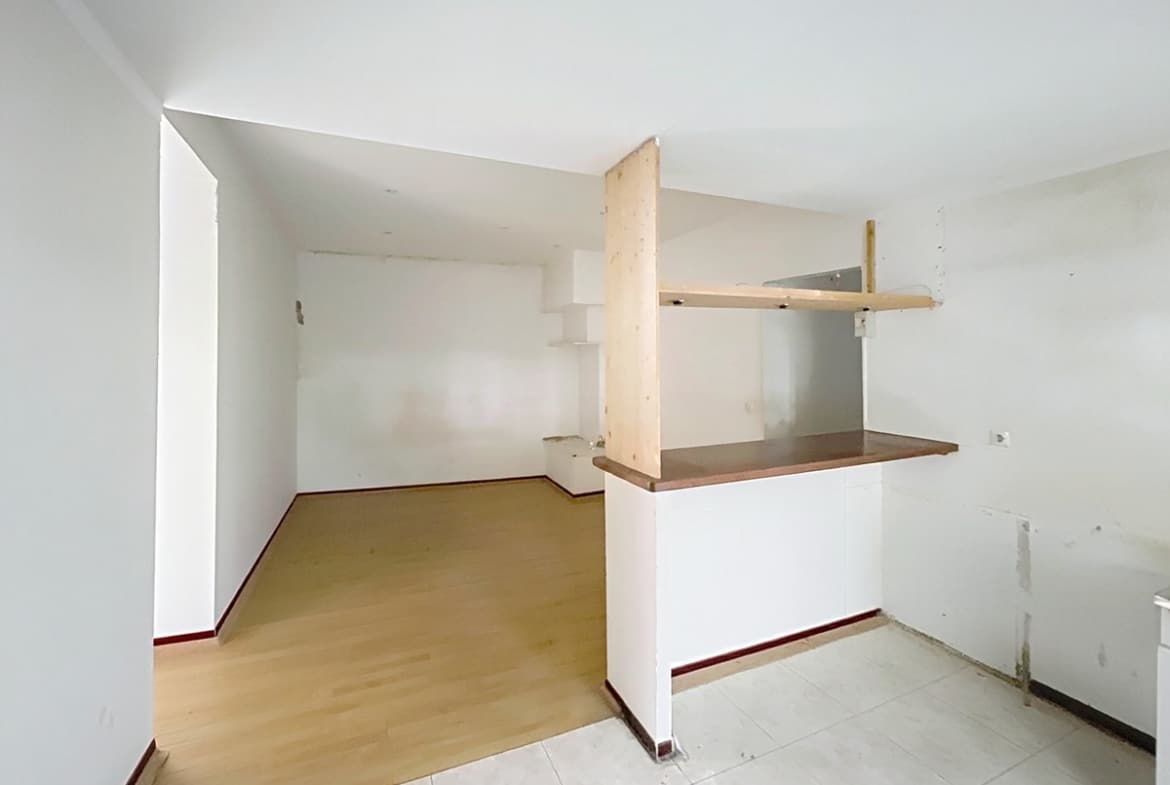 Cuisine avec carrelage blanc au sol séparée du séjour par un comptoir bar blanc surmonté d'étagères en bois brut. Les murs présentent des traces d'anciens équipements et le séjour adjacent dispose d'un parquet stratifié clair.