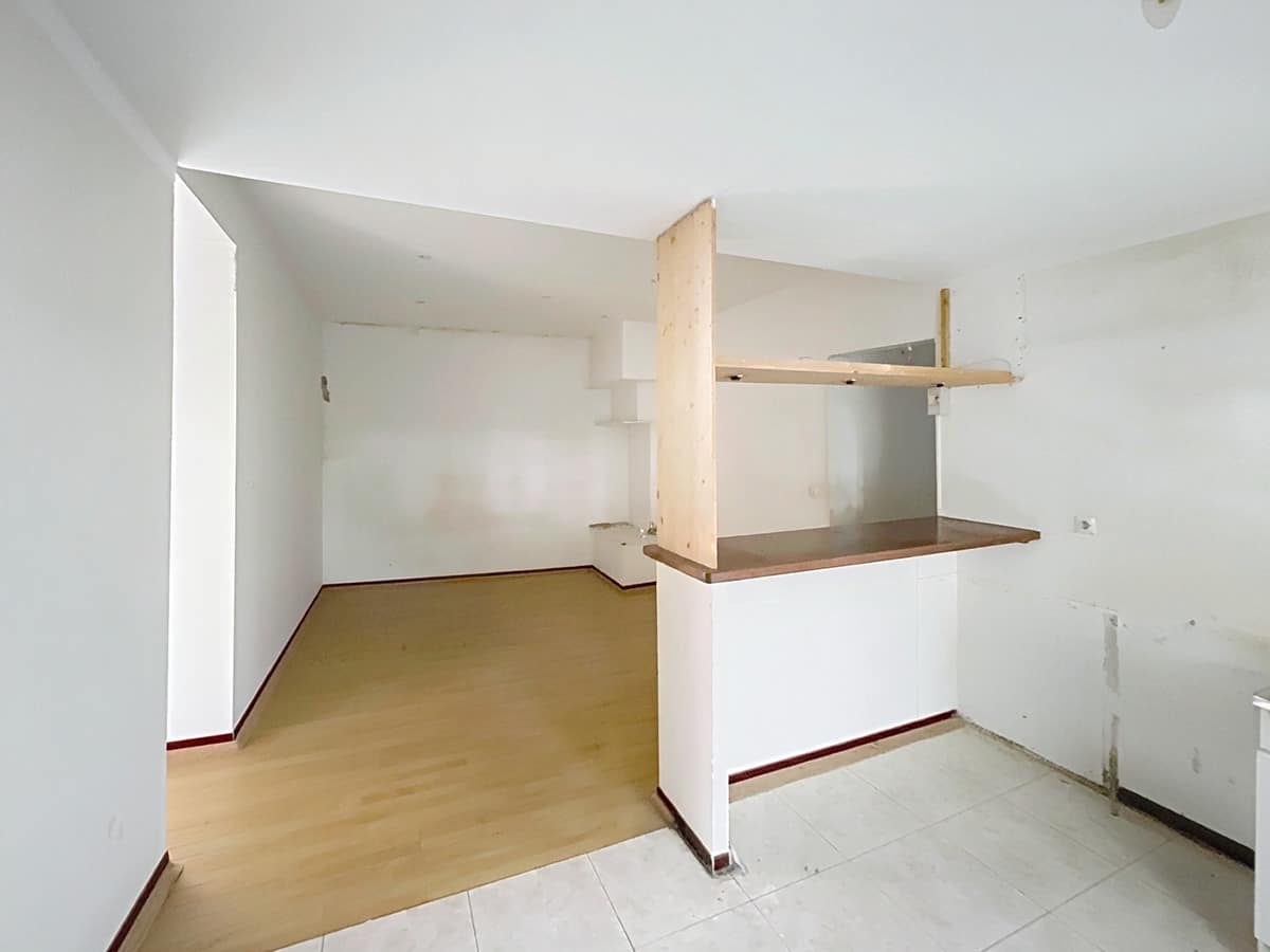 Cuisine avec carrelage blanc au sol séparée du séjour par un comptoir bar blanc surmonté d'étagères en bois brut. Les murs présentent des traces d'anciens équipements et le séjour adjacent dispose d'un parquet stratifié clair.