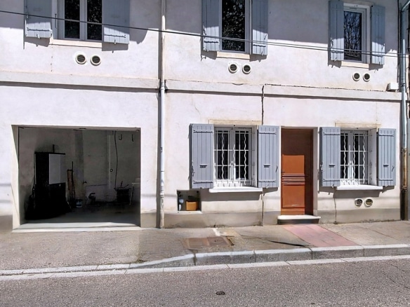 Façade d'une maison de ville à deux niveaux avec enduit clair et volets gris-bleu, présentant quatre fenêtres à l'étage et une porte d'entrée en bois marron au rez-de-chaussée. Un garage ouvert sur la gauche laisse apparaître un espace de rangement, tandis que des grilles de protection équipent les fenêtres du niveau inférieur.