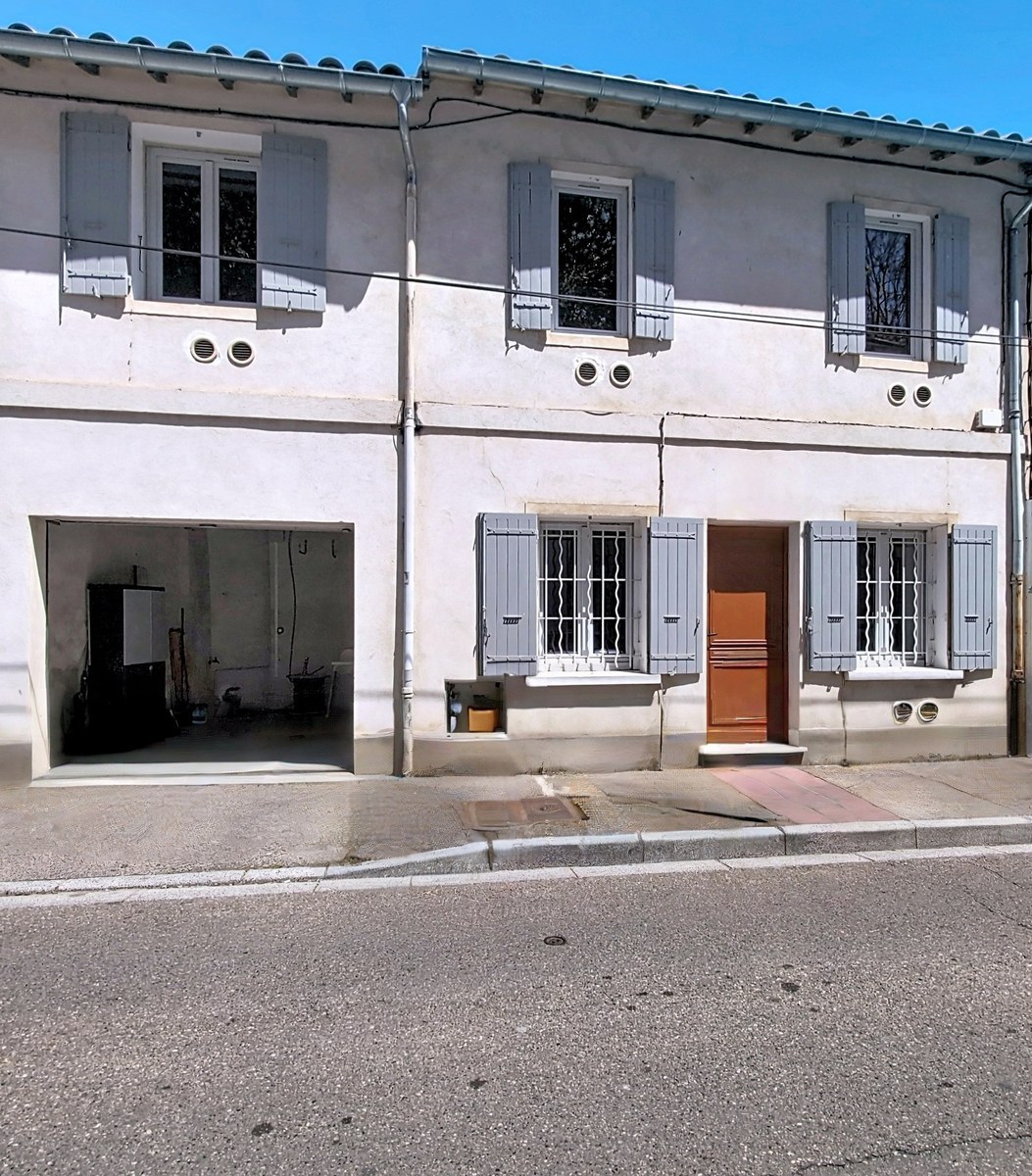 Façade d'une maison de ville à deux niveaux avec enduit clair et volets gris-bleu, présentant quatre fenêtres à l'étage et une porte d'entrée en bois marron au rez-de-chaussée. Un garage ouvert sur la gauche laisse apparaître un espace de rangement, tandis que des grilles de protection équipent les fenêtres du niveau inférieur.