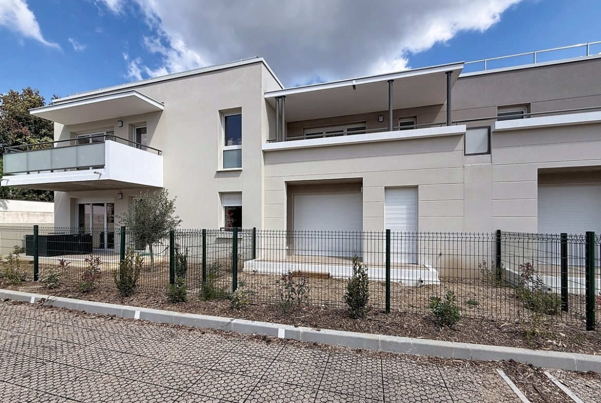 Façade moderne de résidence en crépi beige et gris avec balcons à garde-corps en verre. Jardins privatifs clôturés au rez-de-chaussée avec oliviers, ciel partiellement nuageux en arrière-plan.