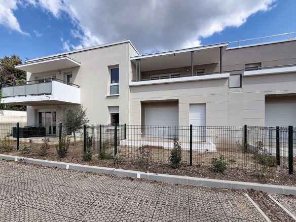 Façade moderne de résidence en crépi beige et gris avec balcons à garde-corps en verre. Jardins privatifs clôturés au rez-de-chaussée avec oliviers, ciel partiellement nuageux en arrière-plan.