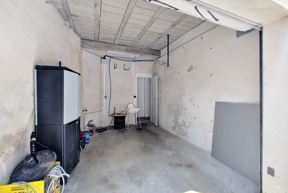 Garage brut avec sol en béton, murs en crépi ancien et plafond avec poutres apparentes. On distingue une armoire de rangement noire, un aspirateur Kärcher jaune et des équipements de bricolage. Une porte blanche donne accès à l'intérieur.
