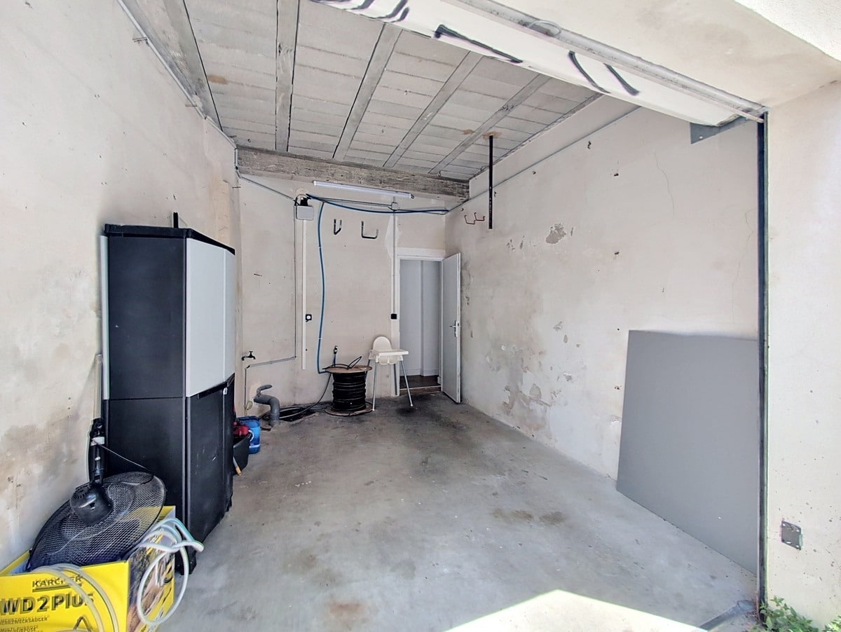 Garage brut avec sol en béton, murs en crépi ancien et plafond avec poutres apparentes. On distingue une armoire de rangement noire, un aspirateur Kärcher jaune et des équipements de bricolage. Une porte blanche donne accès à l'intérieur.