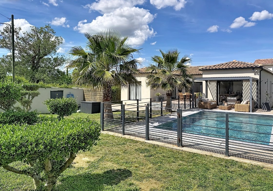 Jardin paysager avec gazon et arbustes taillés devant une piscine rectangulaire entourée d'une clôture métallique noire. Maison de plain-pied aux murs clairs avec toiture en tuiles, terrasse couverte équipée de mobilier en rotin et palmiers en arrière-plan sous un ciel partiellement nuageux.
