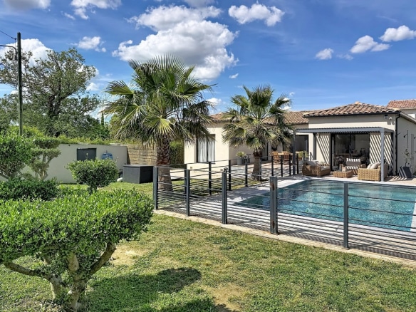 Jardin paysager avec gazon et arbustes taillés devant une piscine rectangulaire entourée d'une clôture métallique noire. Maison de plain-pied aux murs clairs avec toiture en tuiles, terrasse couverte équipée de mobilier en rotin et palmiers en arrière-plan sous un ciel partiellement nuageux.