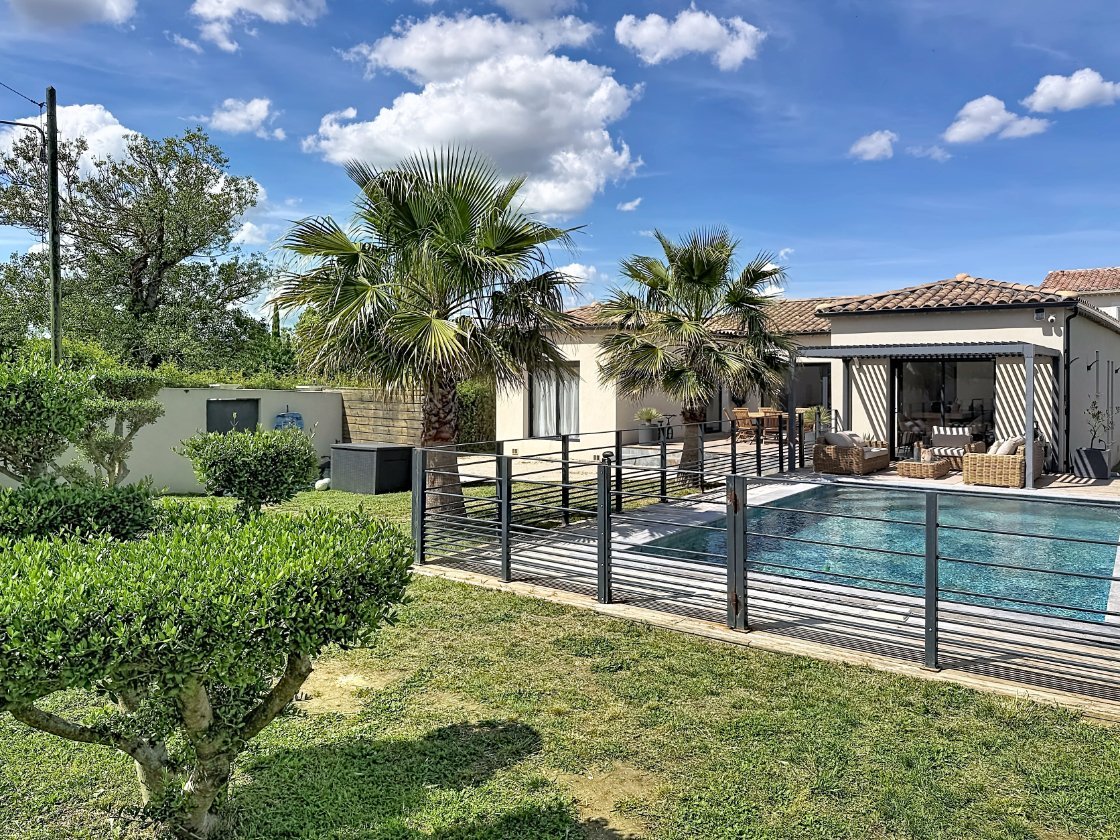 Jardin paysager avec gazon et arbustes taillés devant une piscine rectangulaire entourée d'une clôture métallique noire. Maison de plain-pied aux murs clairs avec toiture en tuiles, terrasse couverte équipée de mobilier en rotin et palmiers en arrière-plan sous un ciel partiellement nuageux.