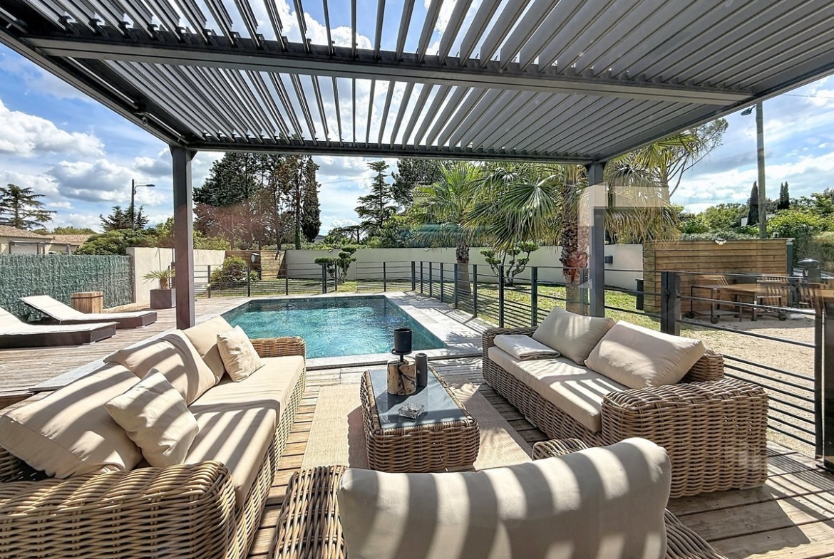 Espace détente sous pergola à lames orientables avec canapés en rotin et coussins beiges à rayures. Piscine rectangulaire à débordement visible au second plan avec transats et jardin paysager avec palmiers.