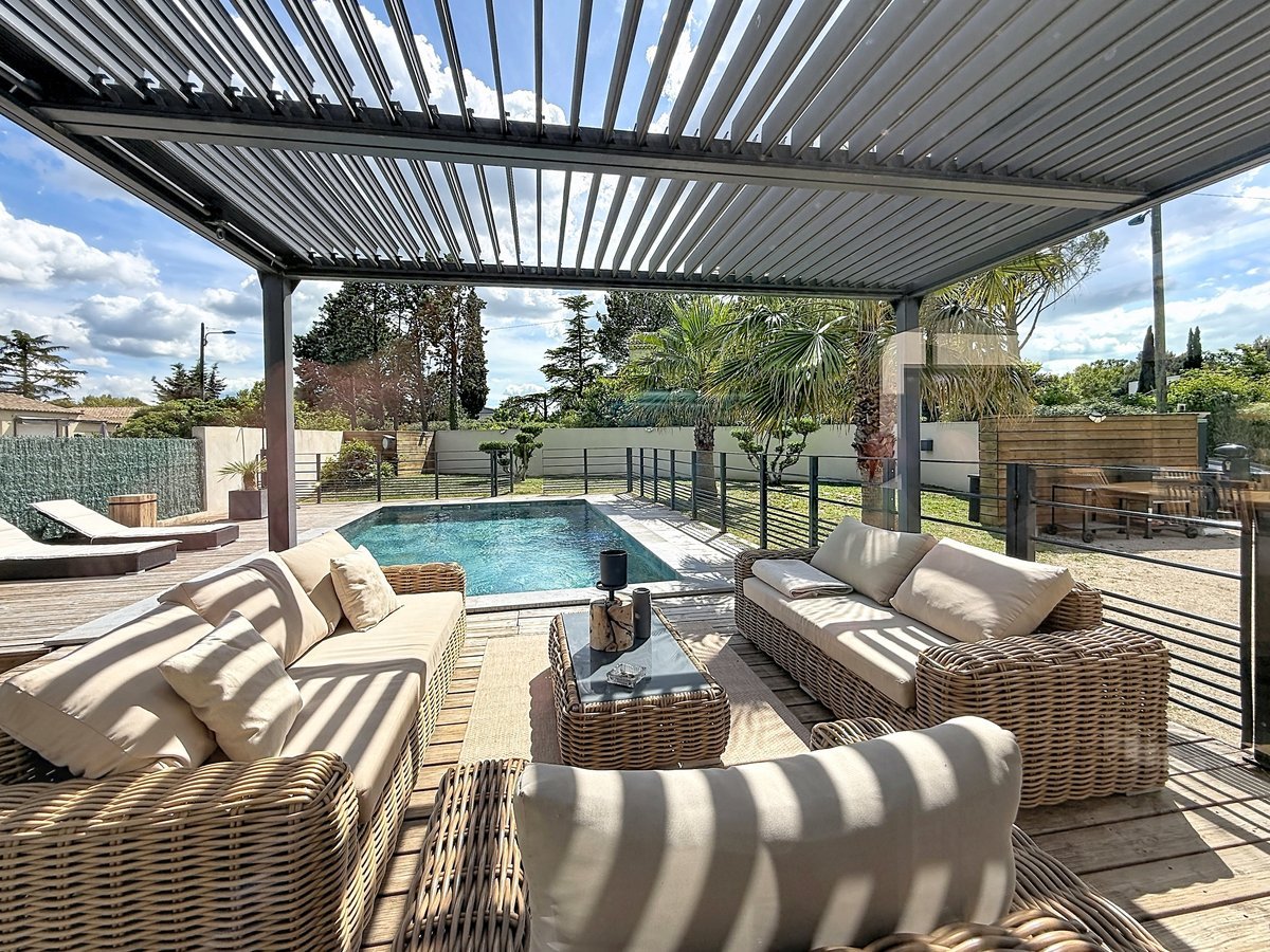 Espace détente sous pergola à lames orientables avec canapés en rotin et coussins beiges à rayures. Piscine rectangulaire à débordement visible au second plan avec transats et jardin paysager avec palmiers.